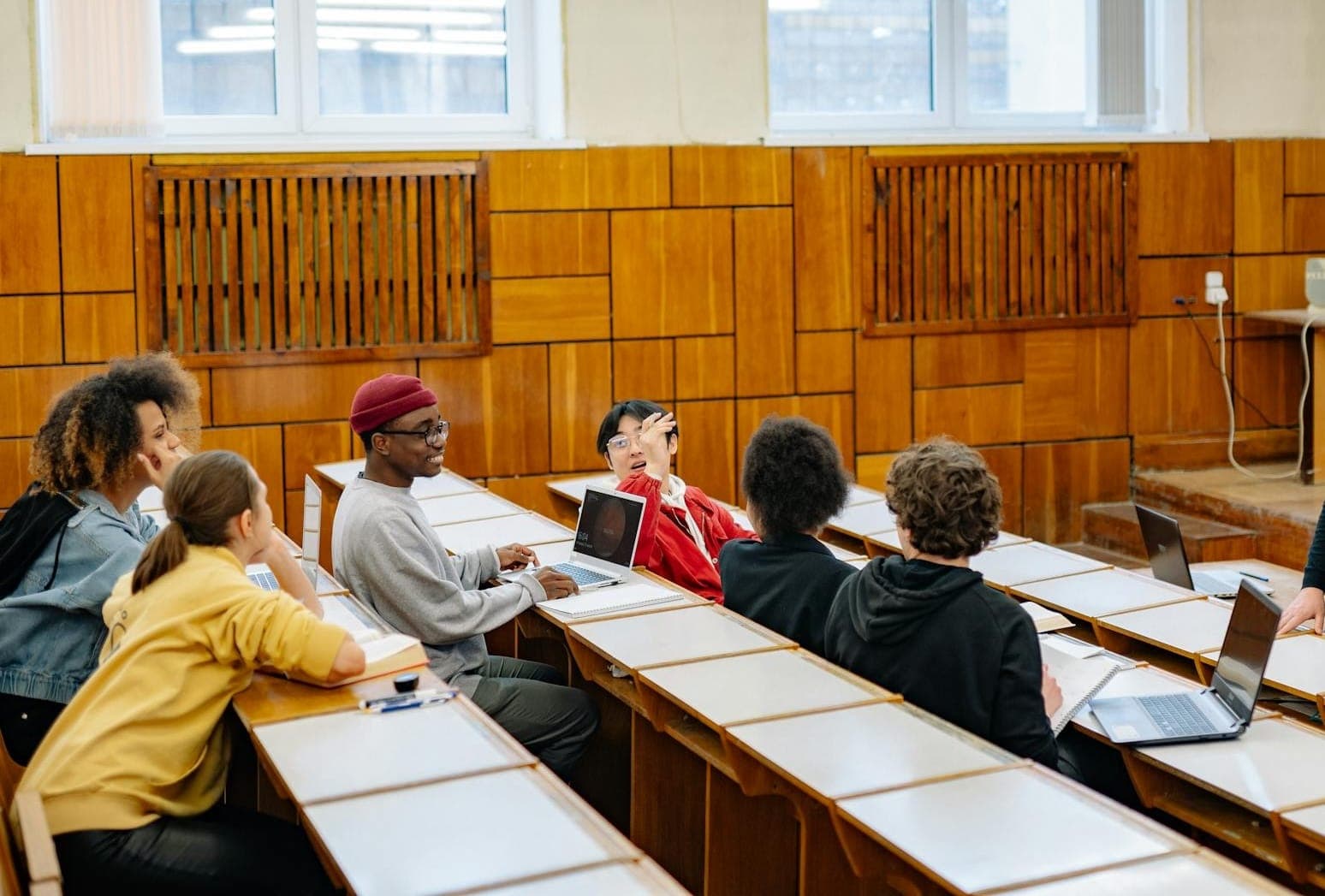 Students sitting in a university lecture hall with laptops and notebooks, with one student raising their hand during a discussion